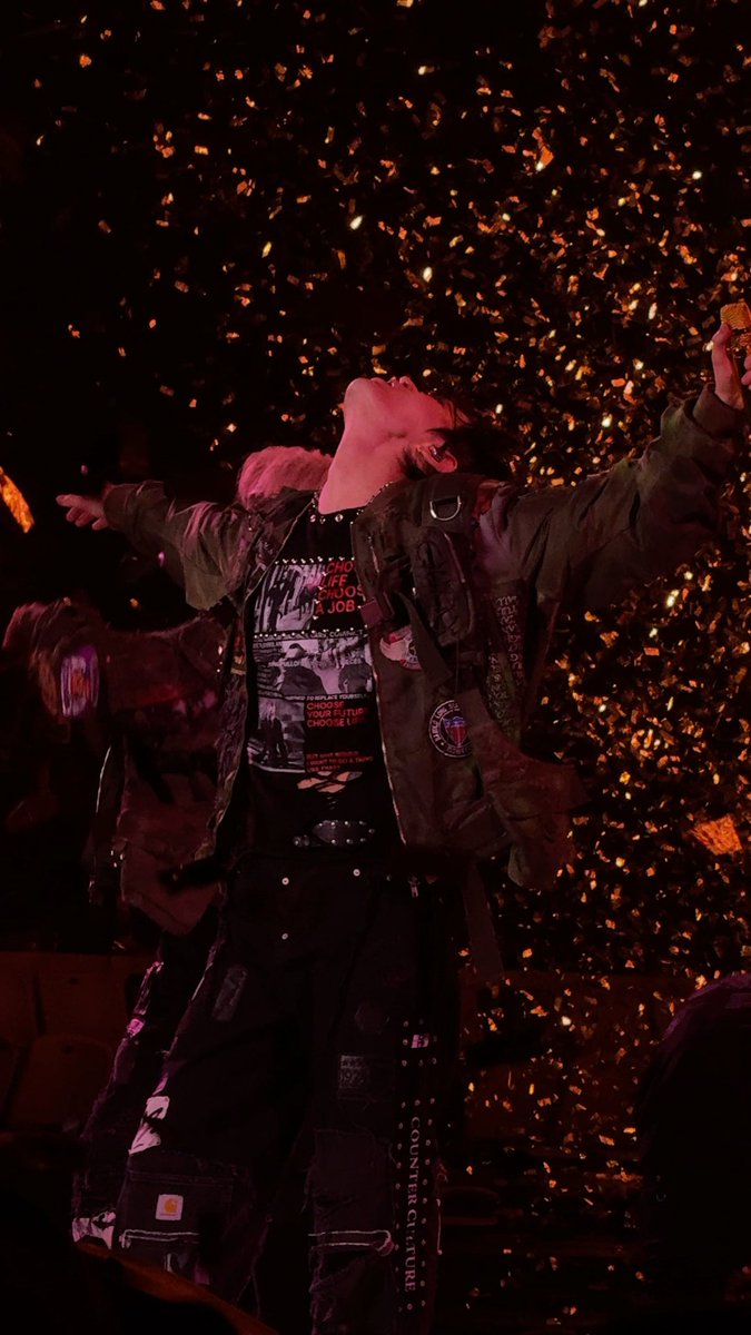A performer with arms outstretched and head tilted back stands on stage amidst a shower of golden confetti, wearing a black graphic tee and an olive jacket adorned with patches. The visible text on the merchandise, including "CHOOSE LIFE" and "COUNTER CULTURE," along with the post's hashtags #YUTA and #NCT127, confirms this is NCT 127 Yuta during THE REDLINE concert in Jakarta.