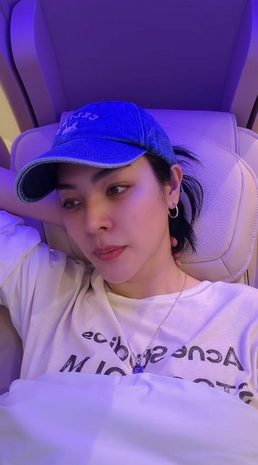 A selfie of a young woman wearing a blue baseball cap and a white t-shirt with reversed text, sitting in what appears to be an airplane seat illuminated with purple lighting.