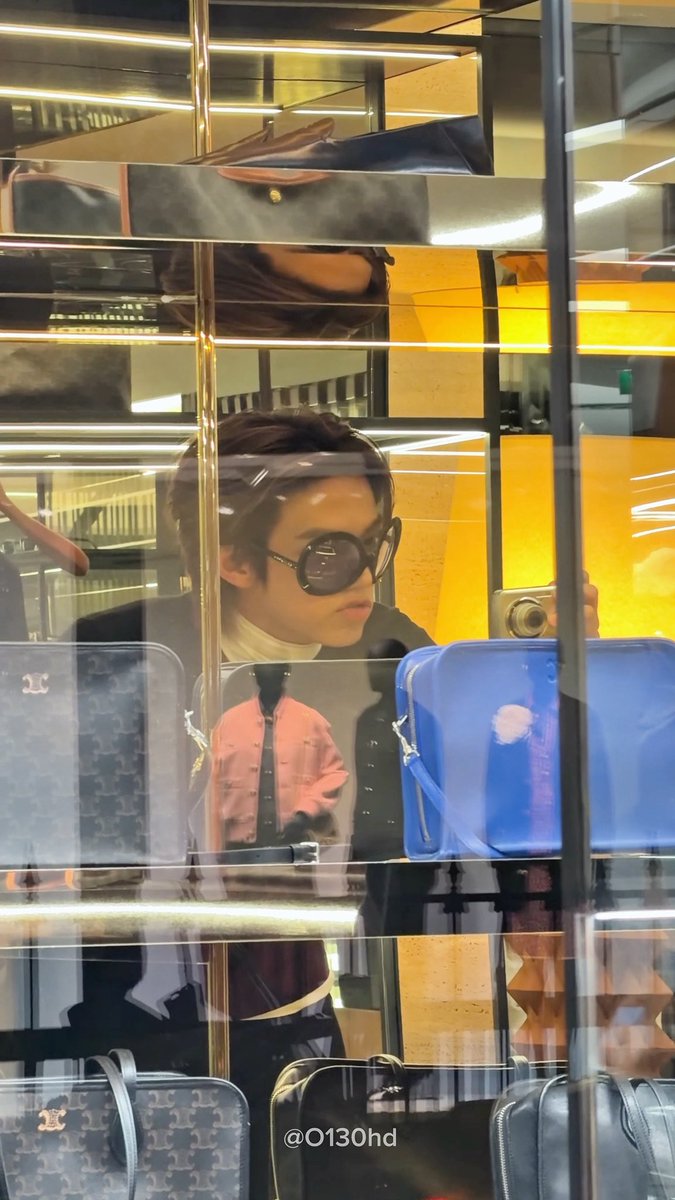 A young man, Dohoon, wearing oversized round sunglasses takes a mirror selfie at a Celine store, surrounded by luxury handbags in glass display cases. The watermark "@O130hd" is visible at the bottom, matching the post's reference to his visit during the TWS x Celine event.