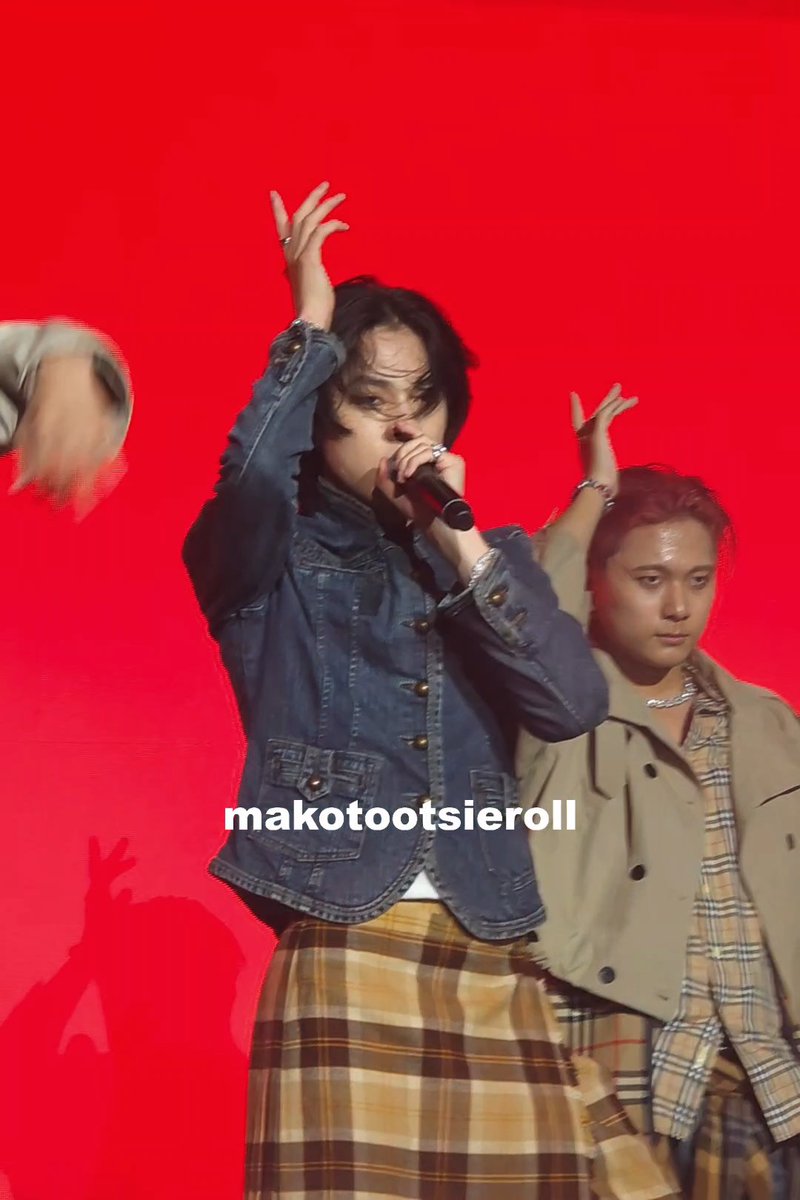 A performer with dark hair sings into a microphone on stage against a bright red background, wearing a denim jacket over a plaid skirt, with the watermark 'makotootsieroll' visible across the image.