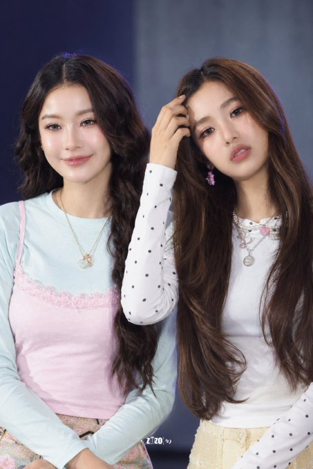 Two young women with long hair pose together in a studio setting; one wears a pastel pink and blue top, while the other wears a white polka-dot shirt and layered necklaces.
