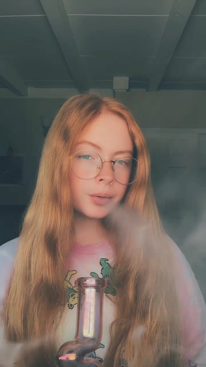 A young woman with long reddish-blonde hair and round wire-frame glasses exhales a cloud of smoke while holding a glass smoking device, dressed in a tie-dye shirt with dinosaur graphics. The hazy, atmospheric photo visually matches the post's reference to her morning "Gas God" smoking session, with the smoke filling the frame alongside the bong.