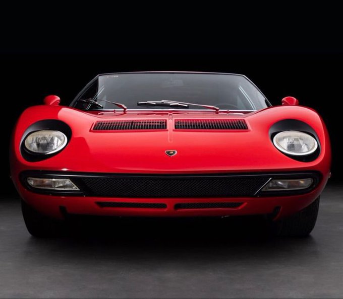 A front view of a classic red Lamborghini Miura sports car displayed against a dark background, featuring its distinctive pop-up headlights and front grille.