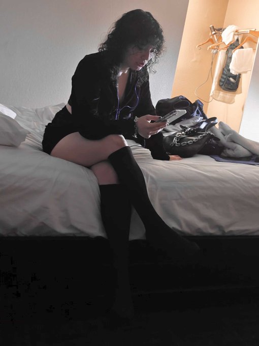 A woman with dark curly hair sitting on a hotel bed, looking down at her phone while wearing a black outfit and thigh-high boots.
