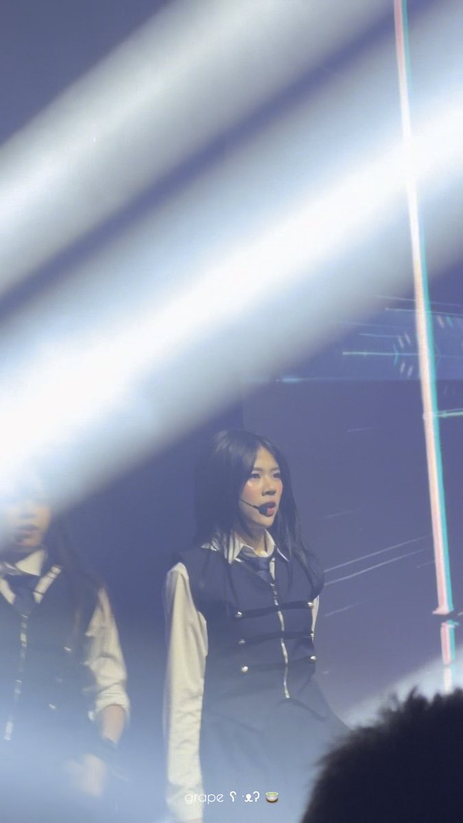 A photo of a K-pop performer on stage with long black hair, wearing a black and white outfit, performing under bright stage lights with another member visible in the background.
