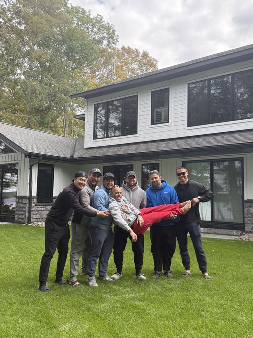 Seven friends gather on a lush lawn in front of a modern home, playfully holding one companion horizontally for the group photo—a joyful moment capturing 25 years of friendship and shared memories, perfectly embodying "The Journey is the Reward."