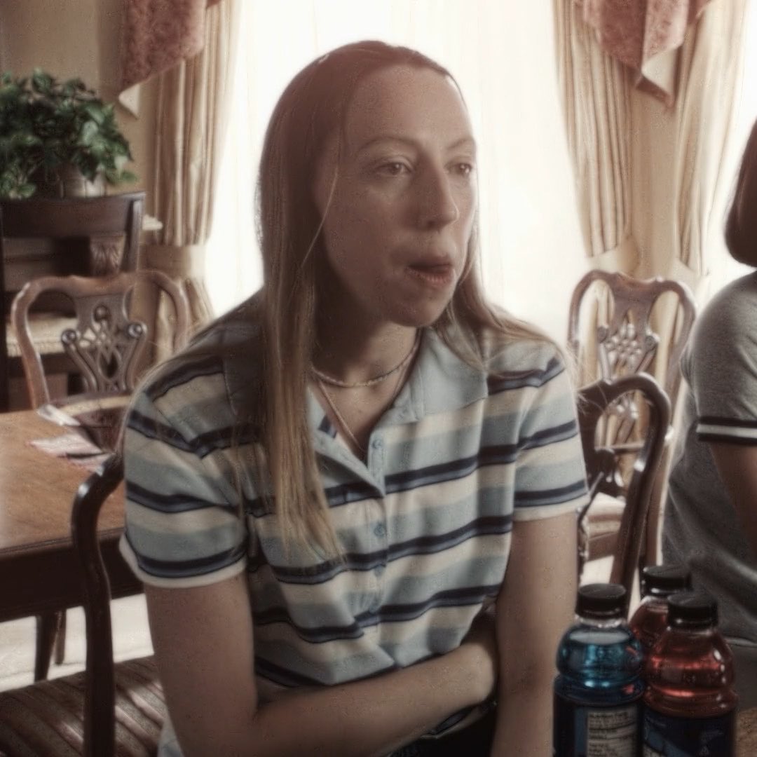 A young woman with long blonde hair wearing a striped polo shirt speaks while sitting at a wooden dining table, with water bottles visible in the foreground.
