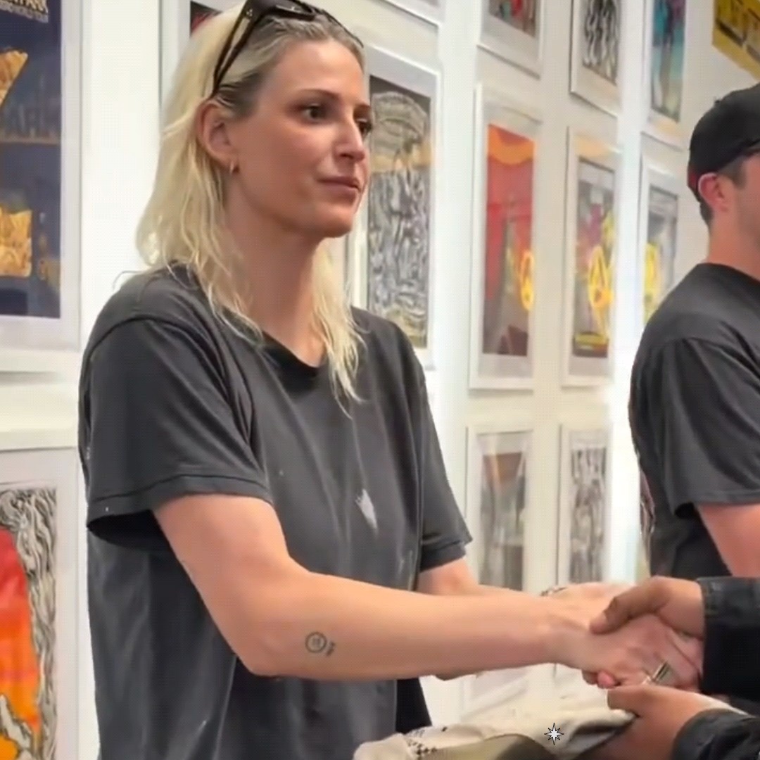 A blonde woman in a dark gray t-shirt stands in a gallery surrounded by framed artwork, shaking hands with someone off-camera while wearing sunglasses on her head.