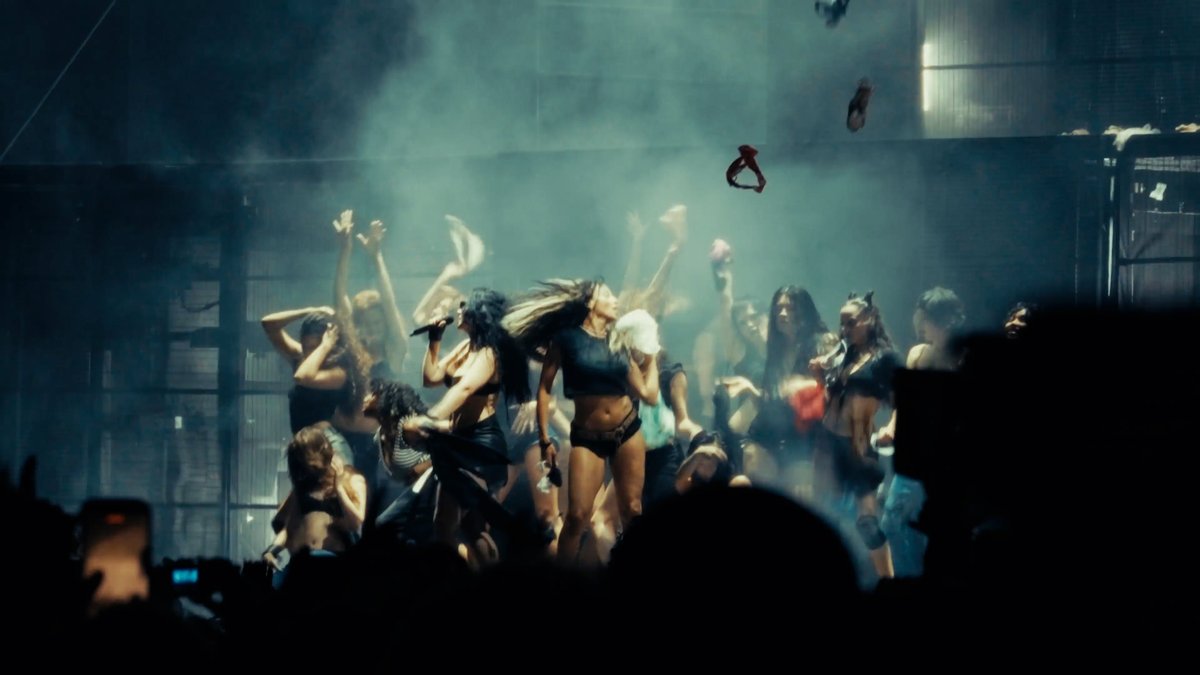 A dynamic concert scene captures a performer singing into a microphone on stage, surrounded by energetic backup dancers with raised arms amid dramatic lighting and haze—visually matching the post's enthusiastic announcement that Day 2 of the event has now opened its gates following a successful Friday.