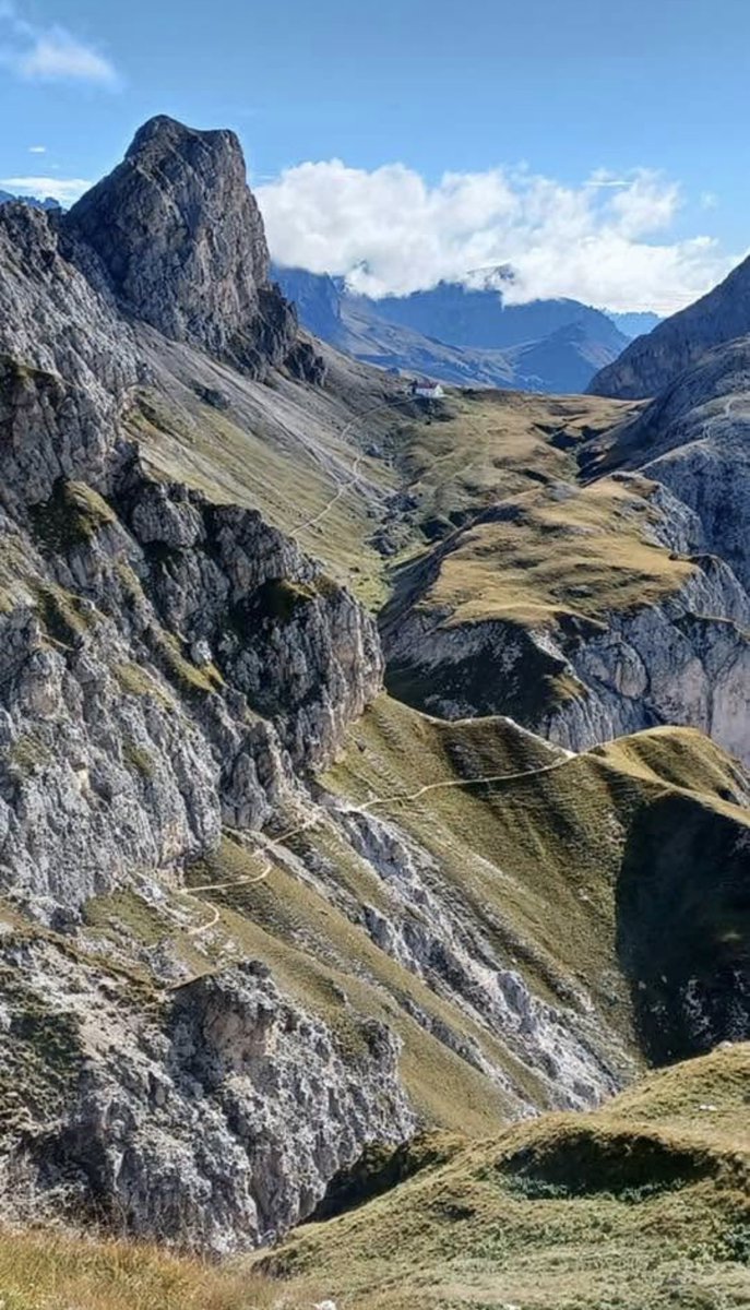 MohoEarth's tweet photo. Ci troviamo sulle Dolomiti, lungo un sentiero escursionistico in quota che attraversa un tipico paesaggio di alta montagna fatto di pareti rocciose calcaree, ghiaioni e valloni alpini.
In lontananza, incastonato nella sella tra i rilievi, si scorge un tipico rifugio alpino che fa da punto di riferimento per gli escursionisti in cammino tra i passi e le cime dolomitiche.