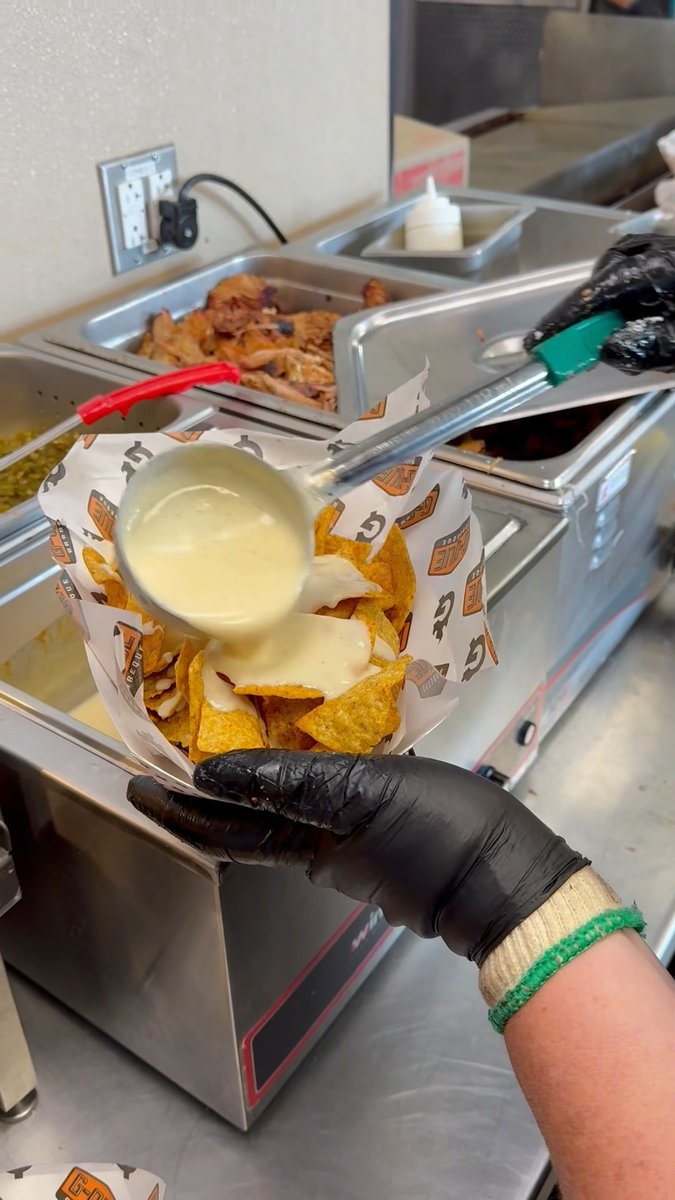 A person wearing a black glove and green wristband uses a ladle to pour creamy white cheese sauce over a basket of tortilla chips at a food counter with metal serving pans.