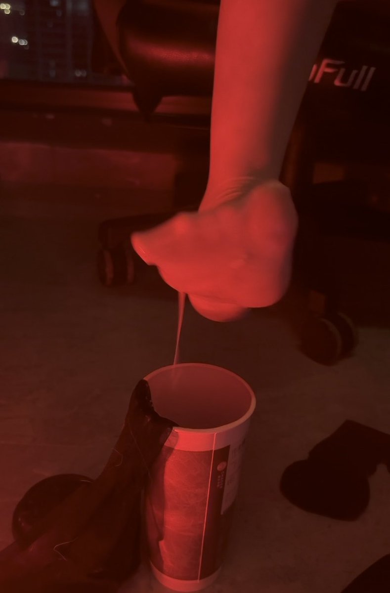 A hand squeezes a thick liquid from a cylindrical container under red lighting, creating a stringy drip in what appears to be a dark indoor setting with an office chair visible in the background.