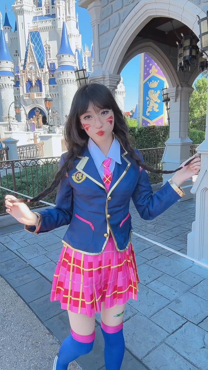 A woman in cosplay with dark hair and face paint poses in front of a Disney castle, wearing a blue blazer, pink plaid skirt, and blue knee-high socks.
