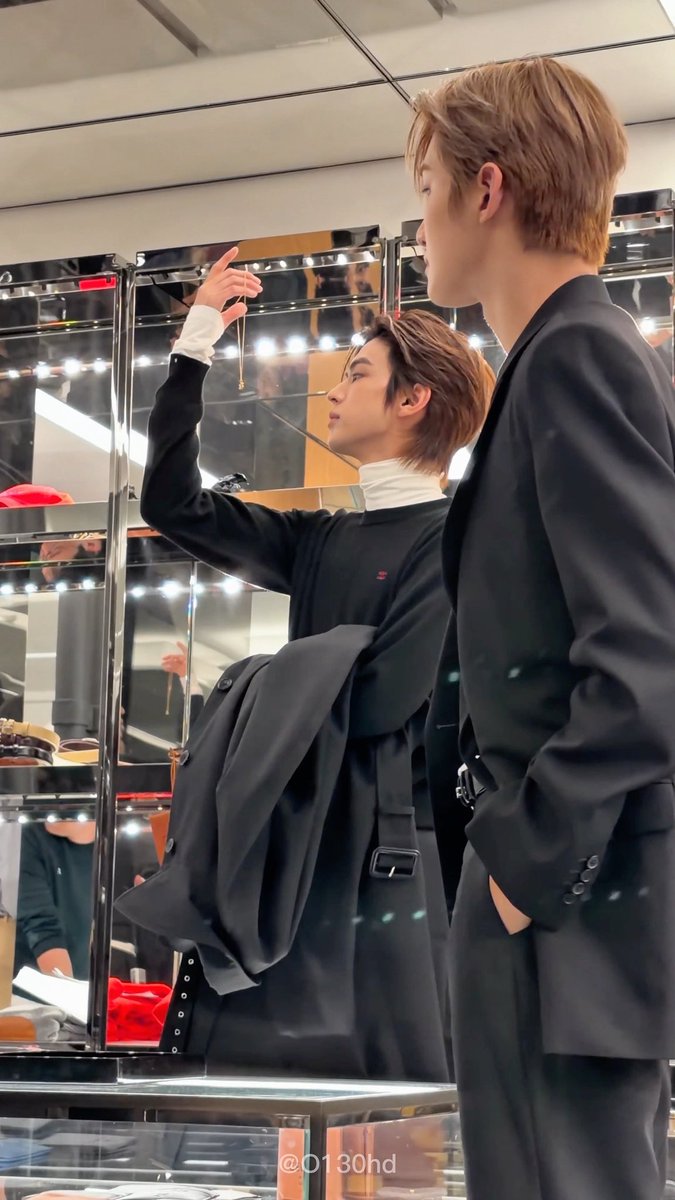 Two young men with brown hair, dressed in black suits, stand in a room with mirrored panels. One holds up a delicate necklace while looking at his reflection, while the other watches him. The watermark reads "@0130hd".