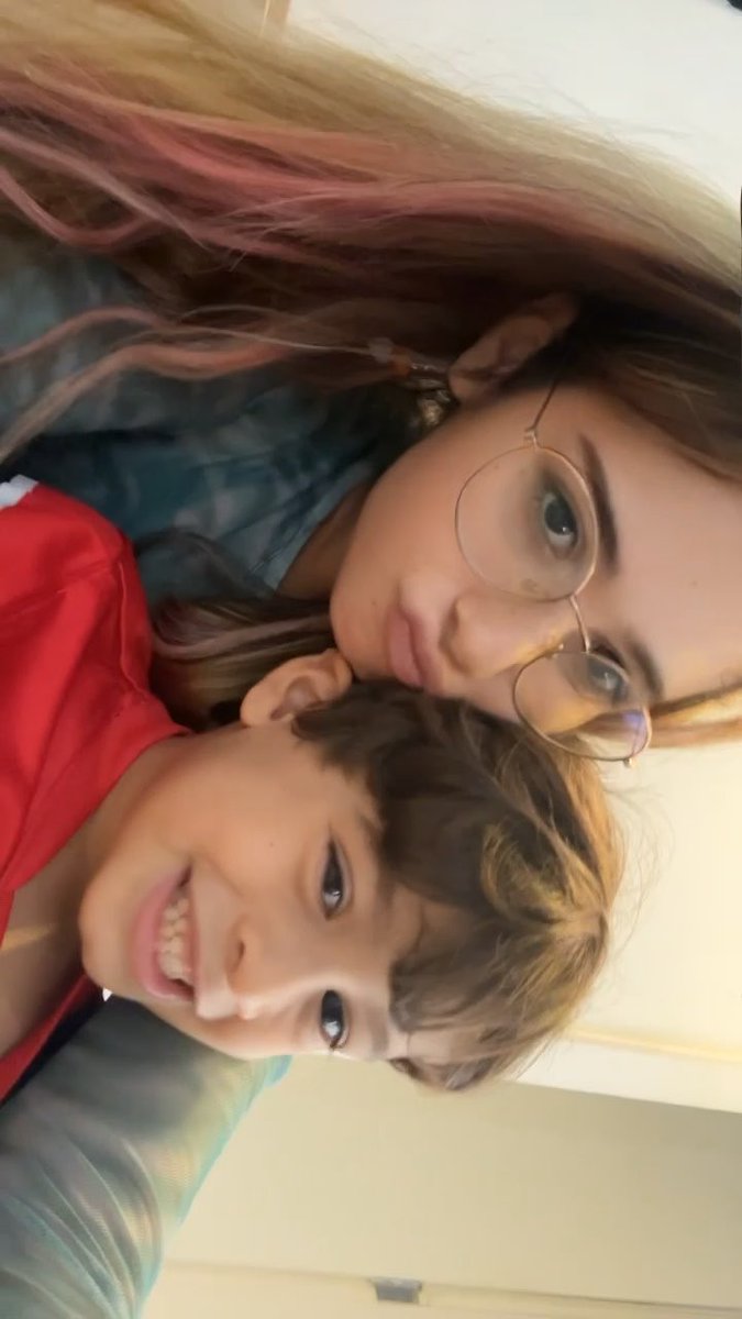 A close-up selfie shows two young people, one with glasses and pink-streaked hair looking calmly at the camera, while the other smiles broadly.