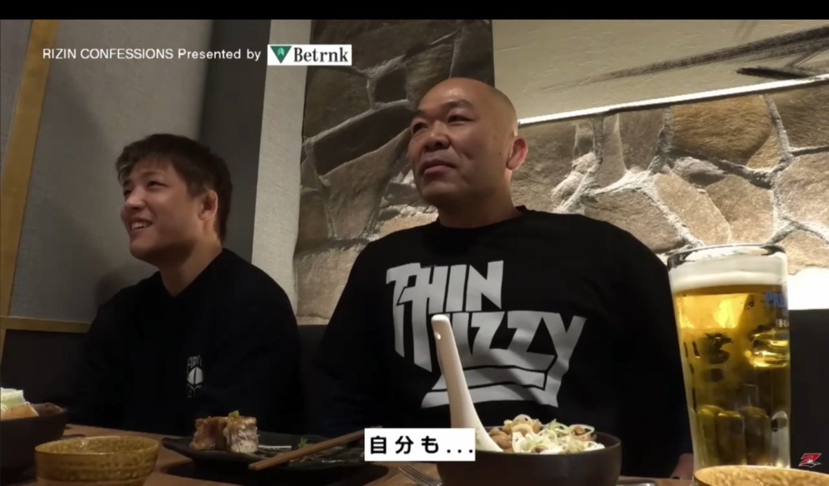 Two men are seated at a restaurant table with beer and food, both smiling and laughing, with the man on the right wearing a black "Thin Lizzy" t-shirt. Japanese subtitles read "自分も..." (meaning "and me too..."), directly referencing the accompanying post about the RIZIN Confessions segment where a participant adds "and me too" to the Horie gay video discussion.