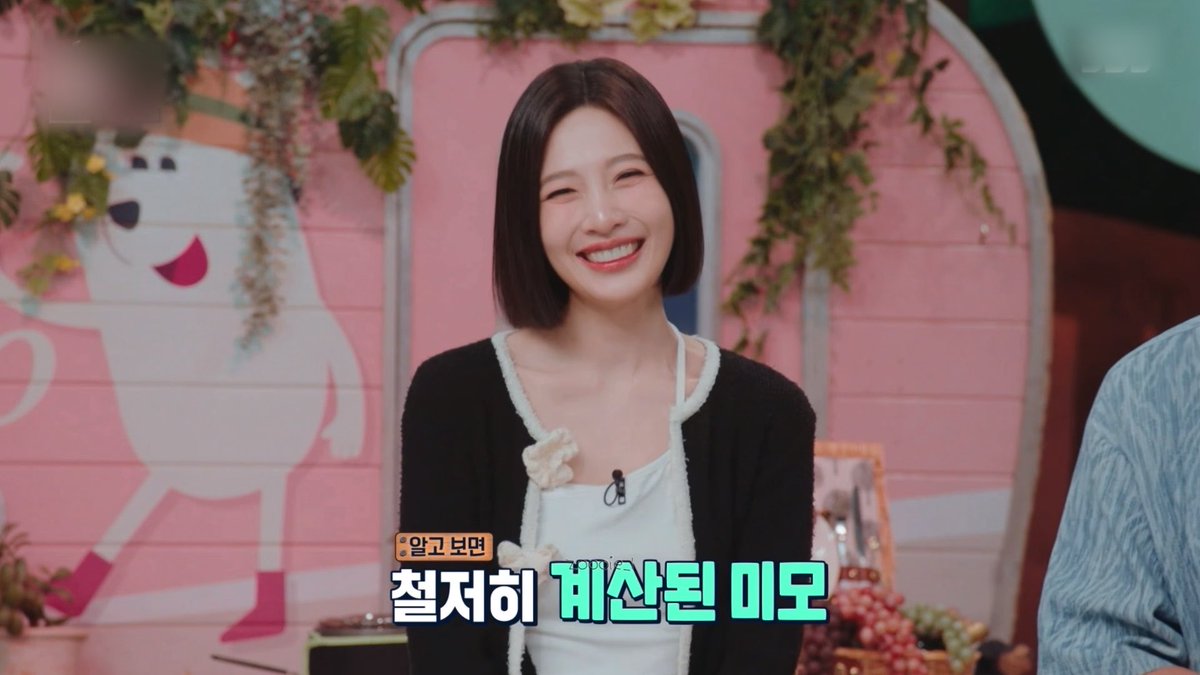 A screenshot from a Korean TV show featuring a smiling woman with short dark hair and red lipstick in a black cardigan over a white top, with Korean subtitles '알고 보면 철저히 계산된 미모' displayed at the bottom.