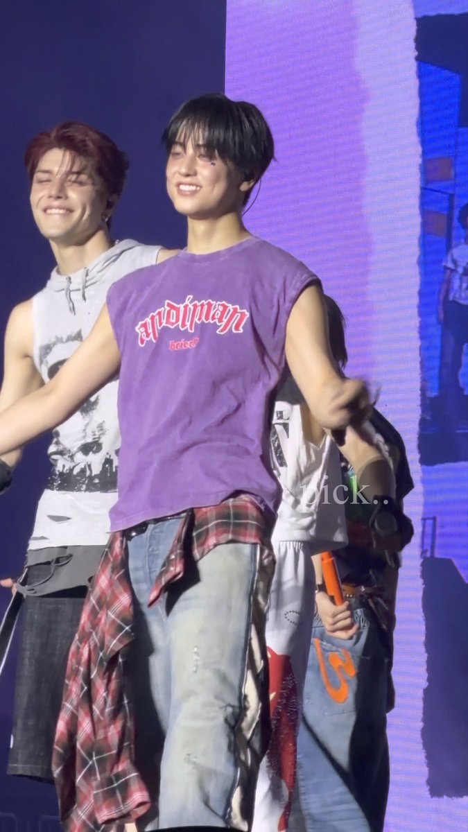 Two young men perform on stage, with one in the foreground wearing a purple sleeveless shirt with red text, smiling, while another man in a white sleeveless top laughs behind him.
