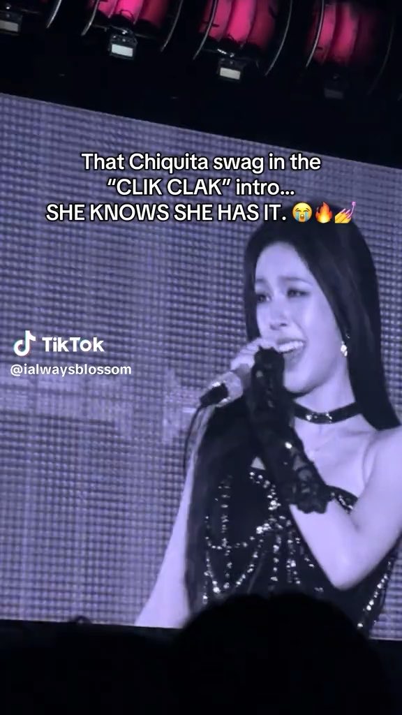 A TikTok screenshot showing a young woman with long dark hair singing into a microphone on a stage with red curtains. The overlaid text reads 'That Chiquita swag in the "CLIK CLAK" intro... SHE KNOWS SHE HAS IT. 😭🔥💅' with the username '@ialwaysblossom'.