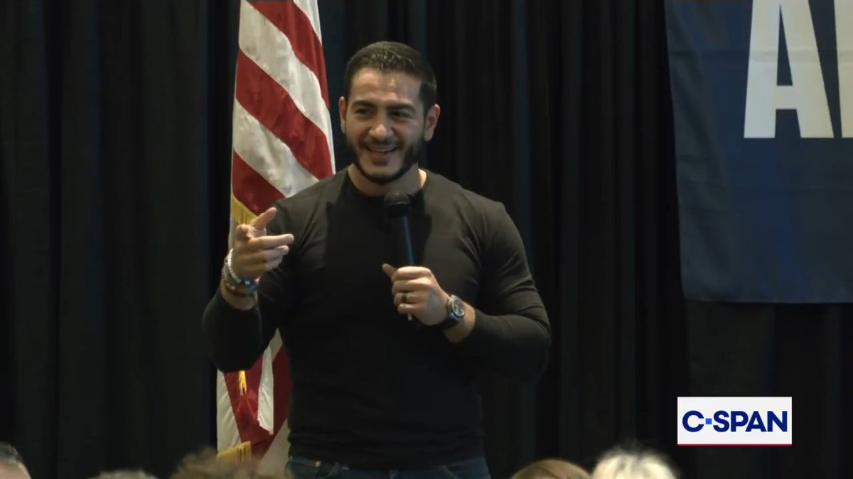 A smiling man with a beard, wearing a black long-sleeve shirt, speaks into a microphone while gesturing with his hand in front of an American flag and a blue banner. The C-SPAN logo appears in the bottom right corner, and the image accompanies a post about Democratic Michigan Senate candidate Abdul El-Sayed criticizing his opponent Mike Rogers during a campaign event.