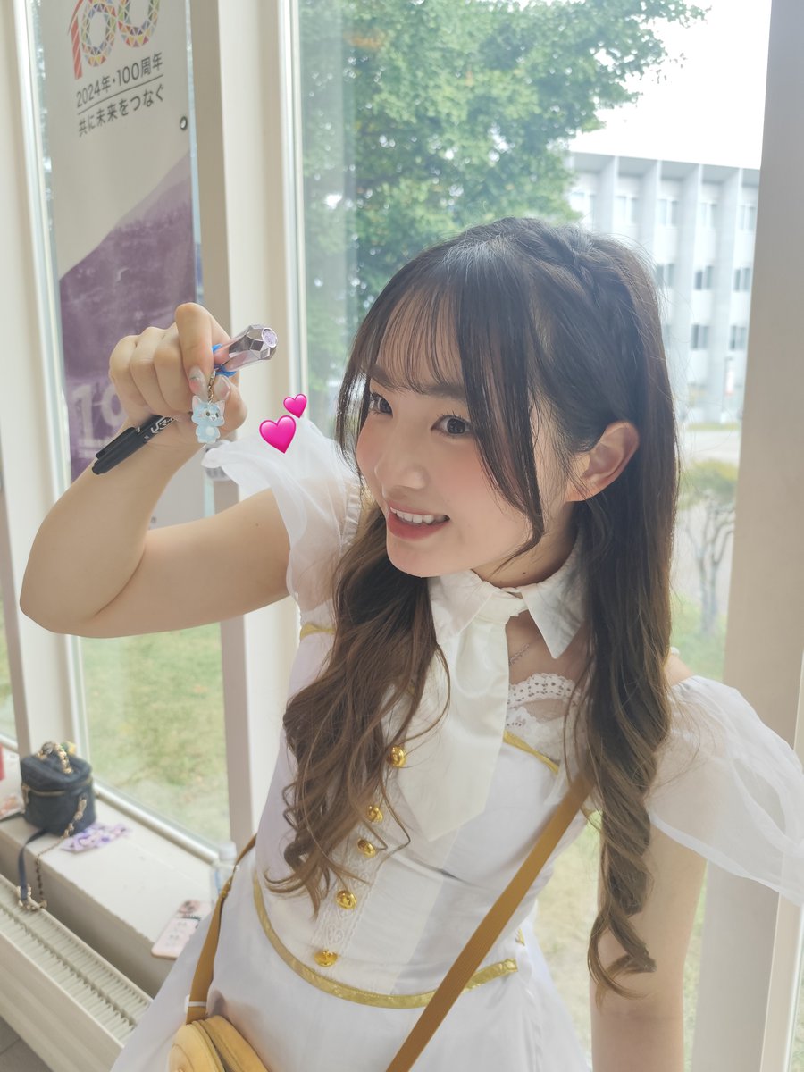 A smiling young woman in a white dress with gold buttons poses holding a pen with a small cat charm, with heart emojis overlaid and a banner reading '2024年・100周年 共に未来をつなぐ' in the background.