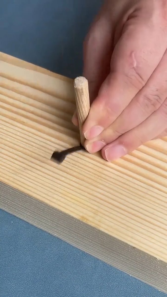 A close-up of a hand using a wooden dowel and a small dark tool to mark or score a groove into a piece of light-colored wood.