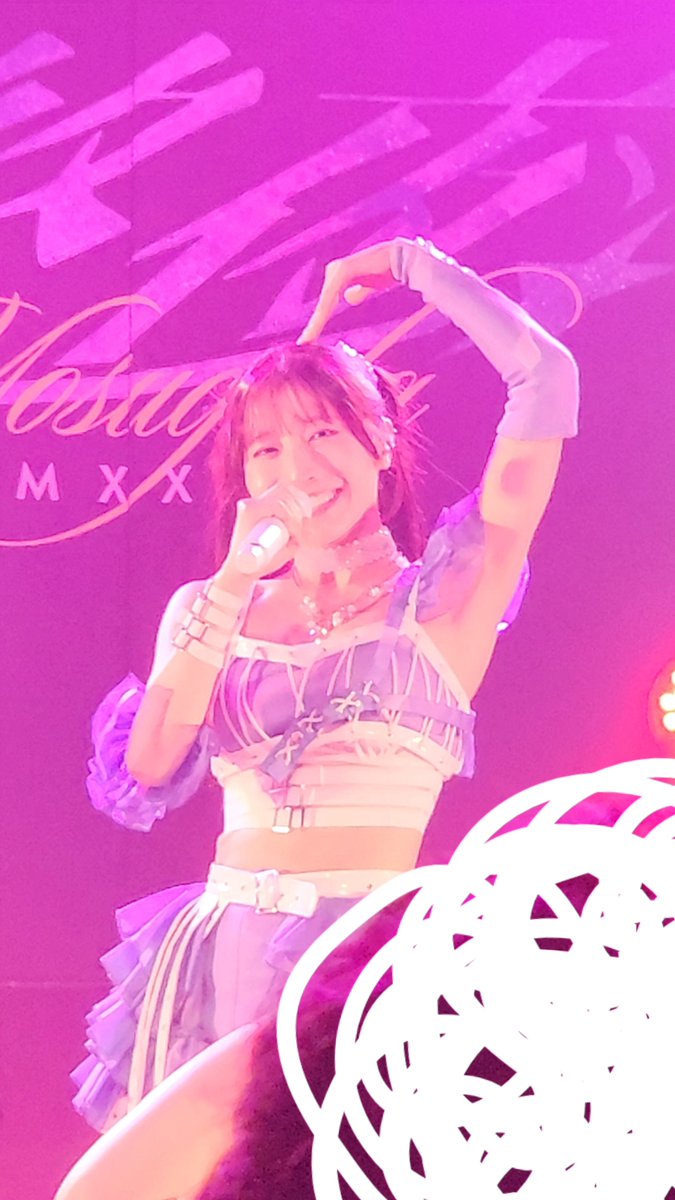 A female performer in a purple and white outfit making a heart shape with her arm while holding a microphone, performing on stage under pink lighting with "Testa RMX" visible on the backdrop.