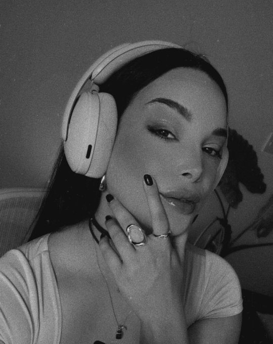A black-and-white selfie of a woman wearing large white over-ear headphones, with dark nail polish and multiple rings on her fingers, posing with her hand near her face.