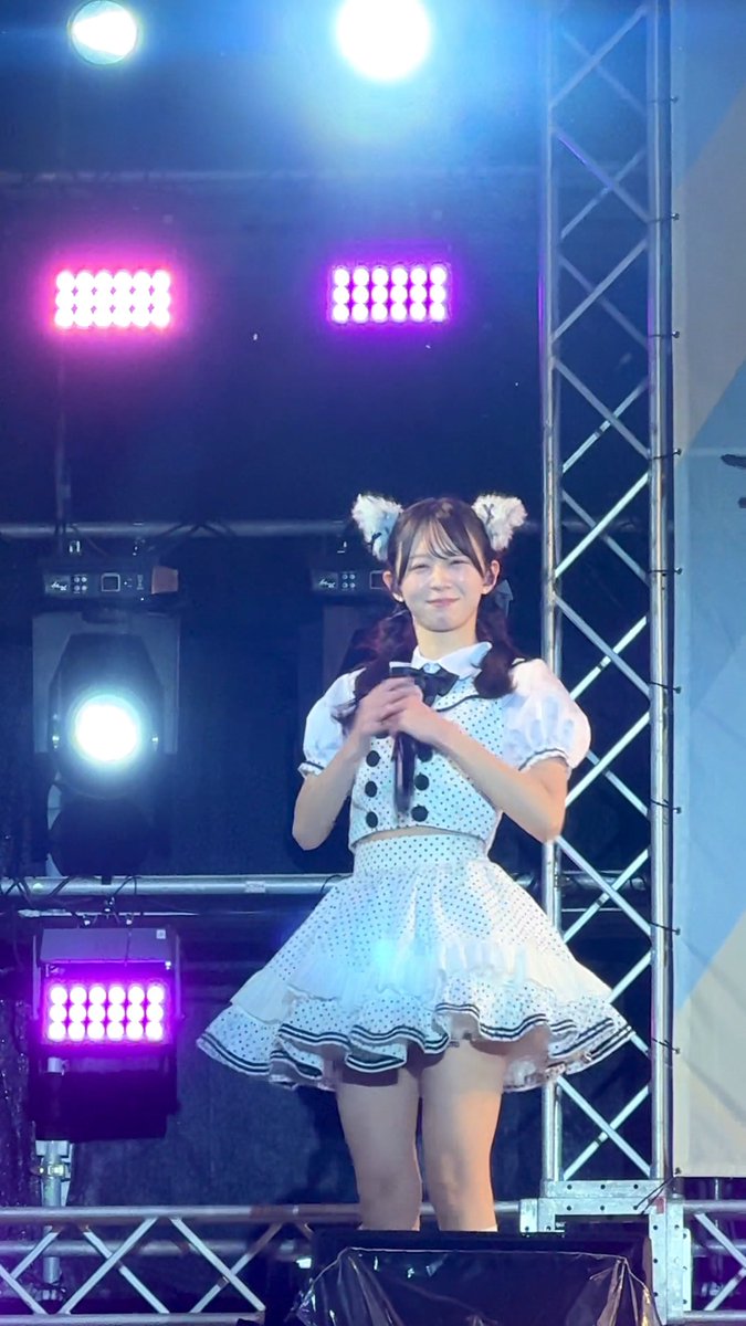 A young Japanese woman wearing a white polka-dot idol costume with cat-ear accessories stands on a stage illuminated by bright spotlights and pink stage lights.