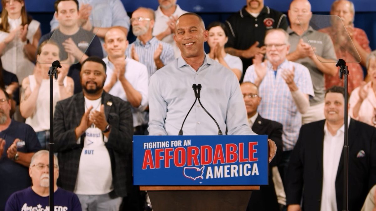 A man stands at a podium speaking to a crowd, with the podium displaying a 'FIGHTING FOR AN AFFORDABLE AMERICA' sign, while audience members applaud behind clear protective shields.