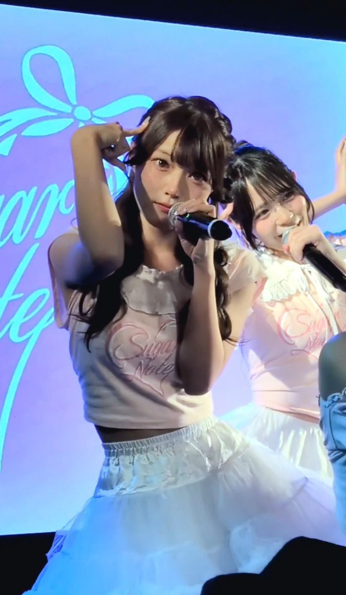 Two female performers in pink tops with 'Sugar Note' logos and white skirts perform on stage, one holding a microphone and making a peace sign.