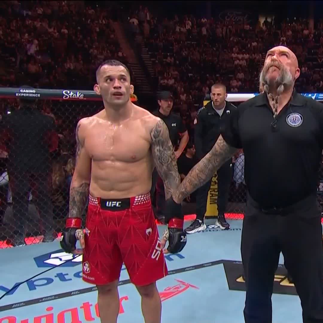 A UFC fighter in red shorts stands inside the octagon next to an official with a distinctive long mustache and braided beard, looking upwards with an emotional expression.