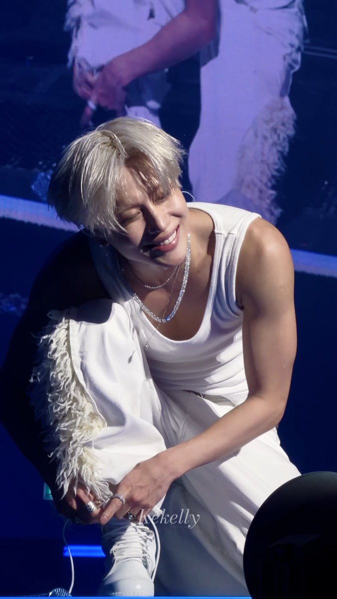 A performer with blonde hair, wearing a white tank top, white pants, and layered silver necklaces, crouches on stage smiling with eyes closed. The image has a watermark that reads 'Kekelly'.
