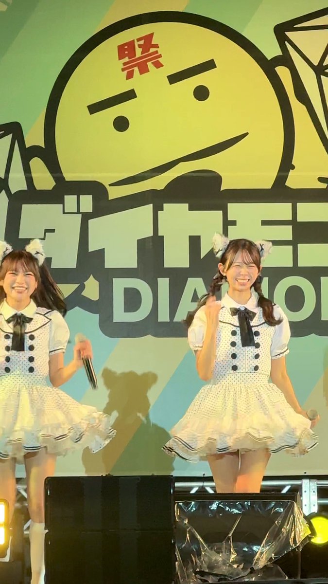 Two smiling performers in white polka-dot dresses with cat ear headbands dance on stage, with Japanese text and 'DIAMOND' visible on the green and blue backdrop.