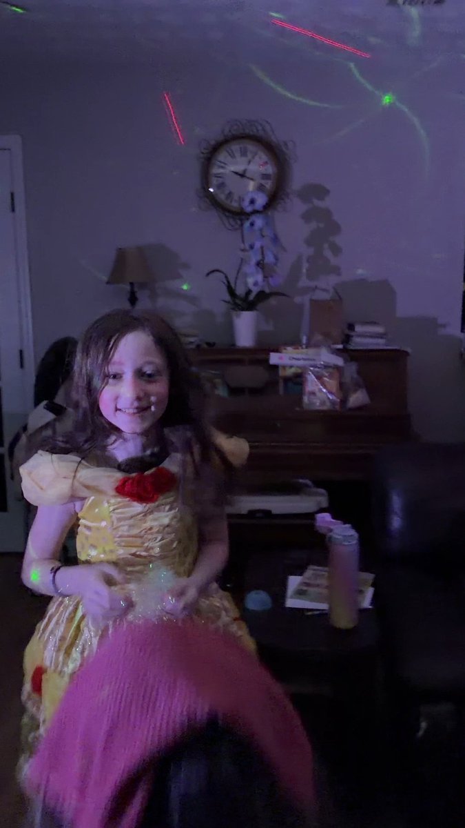 A young girl in a yellow Belle dress smiles while holding a pink fuzzy item at a party, with colorful party lights projected on the wall behind her.