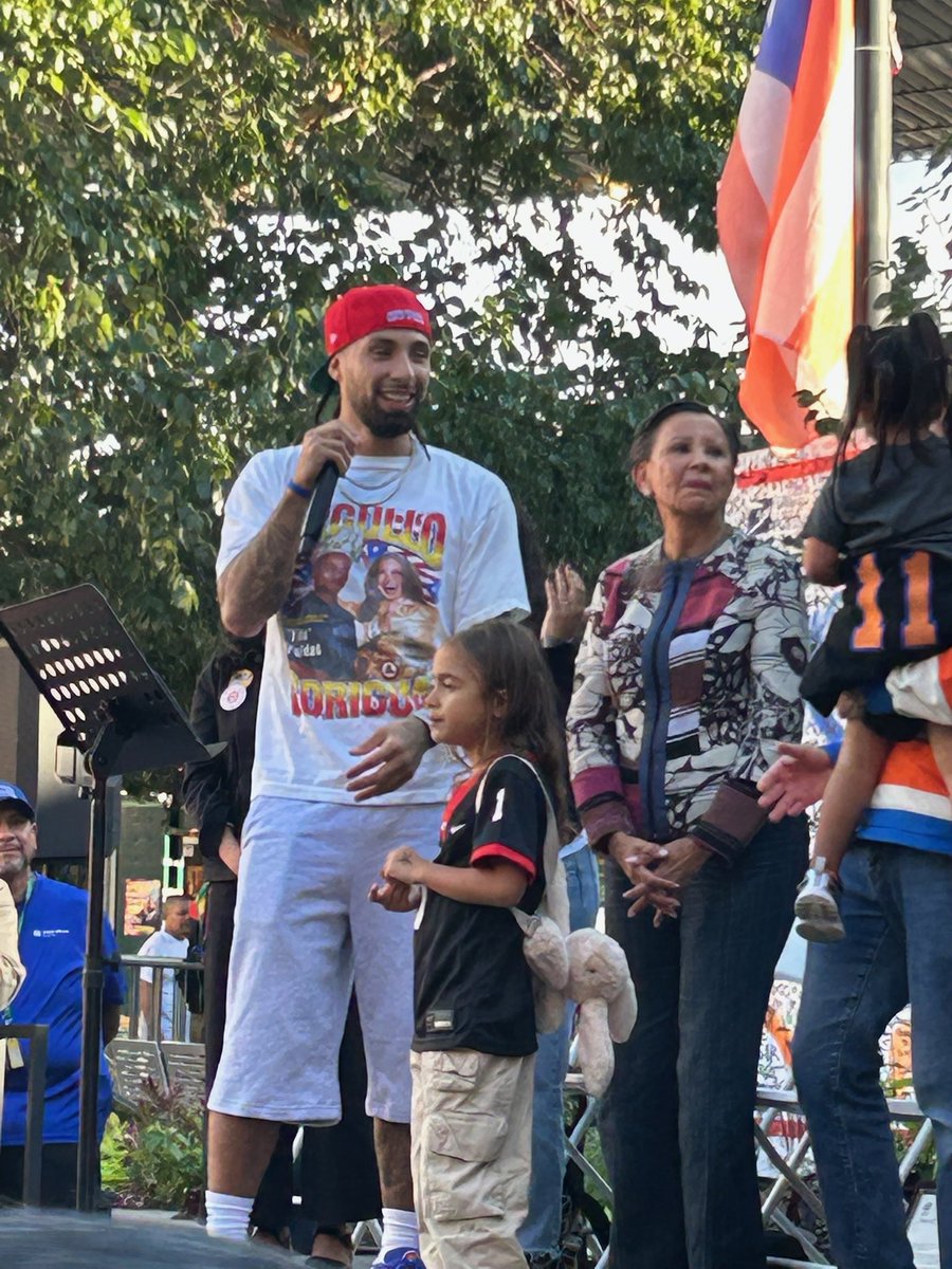 An outdoor gathering on a small stage. A smiling man in a red cap holds a microphone beside a young girl, while a woman and other people stand nearby. Trees and a large red, white, and blue flag are in the background. Some clothing and a display contain text.