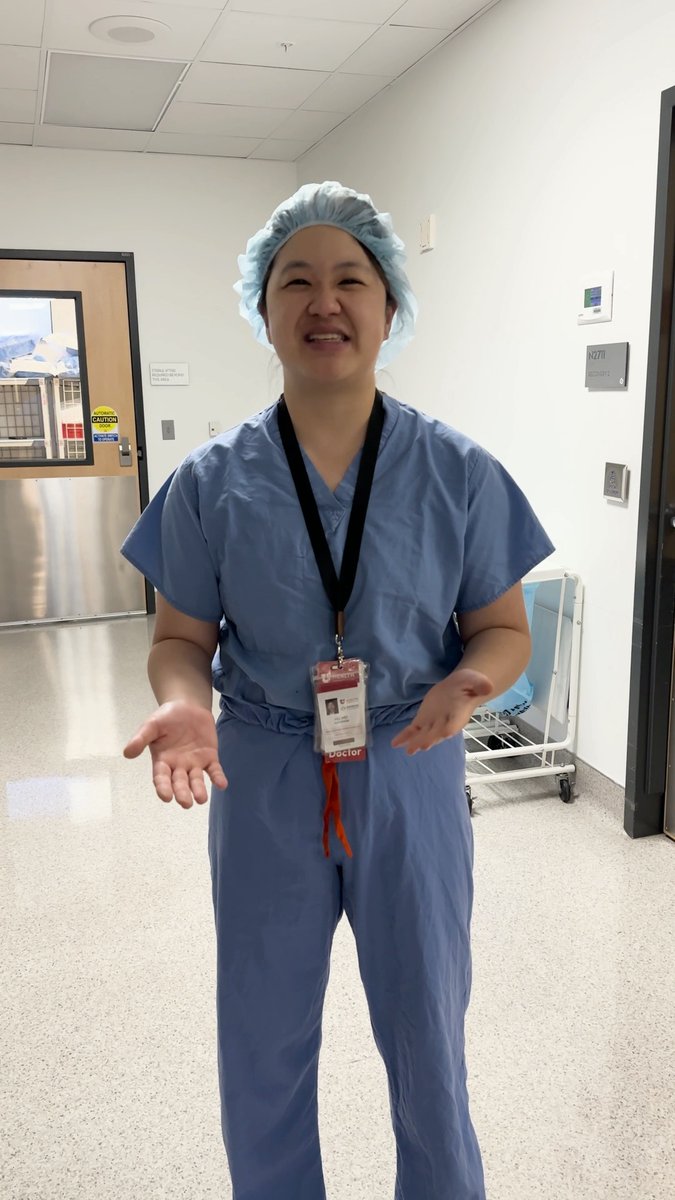 A smiling person wearing blue surgical scrubs and a light blue bouffant cap stands in a hospital hallway, wearing a lanyard with a 'Doctor' ID badge.
