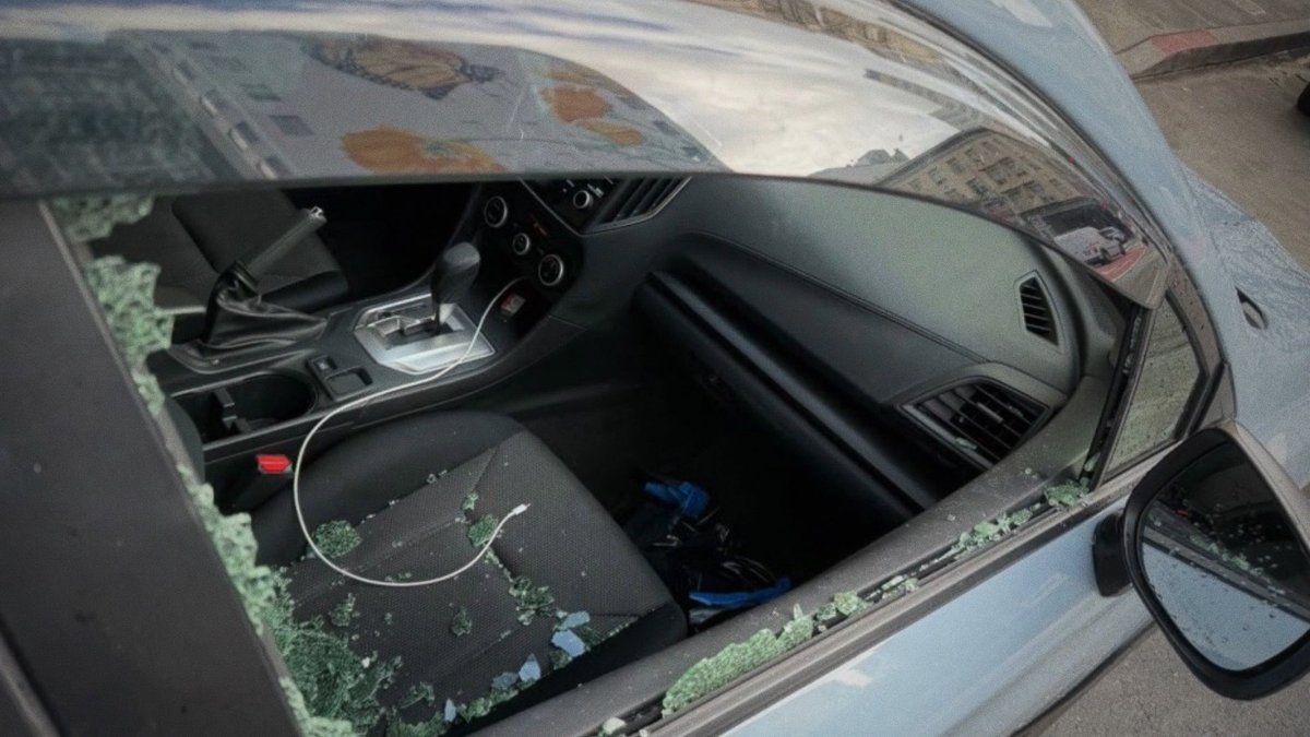 A photo shows a blue car with a shattered driver's side window, glass fragments scattered across the seat, and a charging cable draped over the interior.