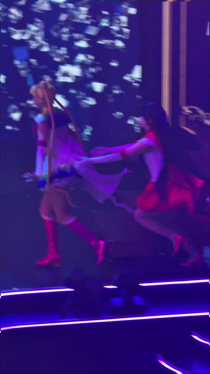 Blurry, dark photograph of two anime-styled performers on stage under purple lighting, appearing to be from a live concert or stage production.