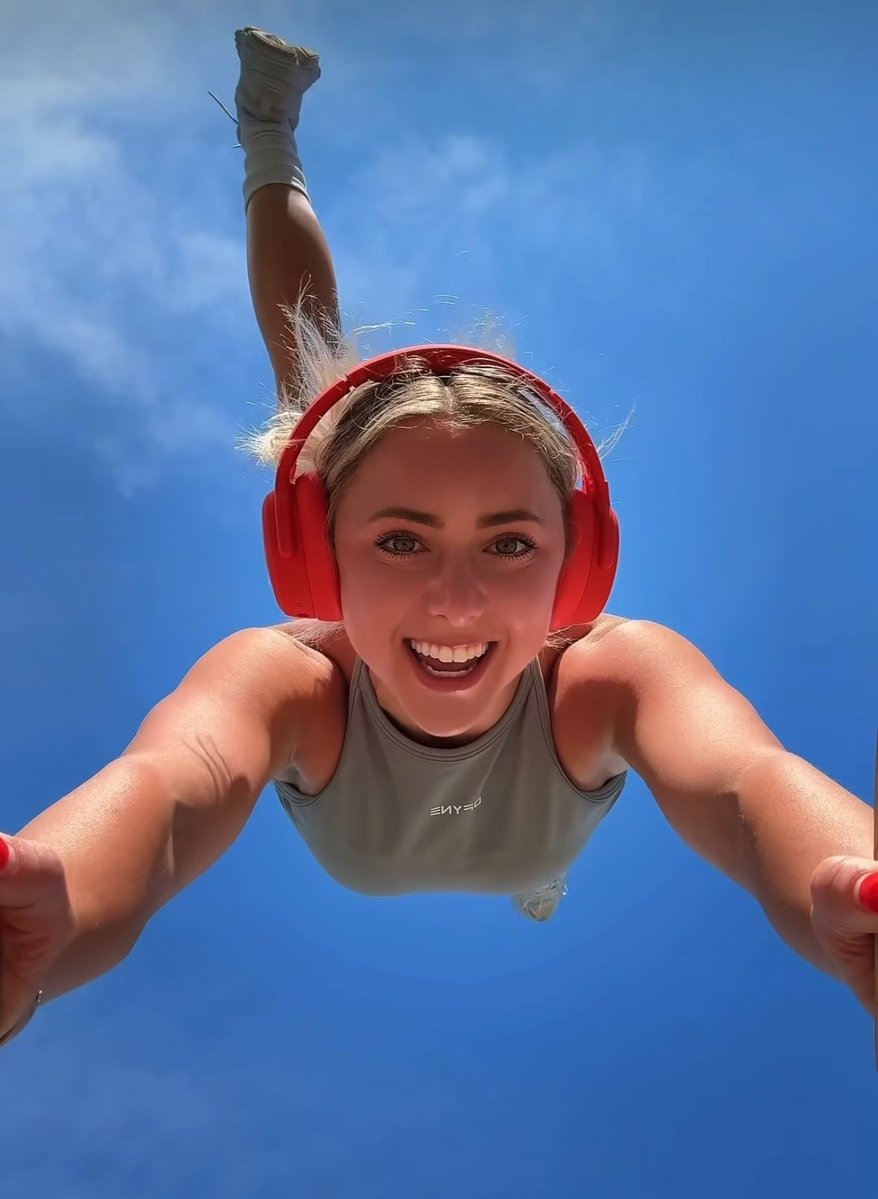 A low-angle selfie of a smiling woman wearing red headphones and a grey tank top, appearing to be in mid-air or doing a handstand against a blue sky background.