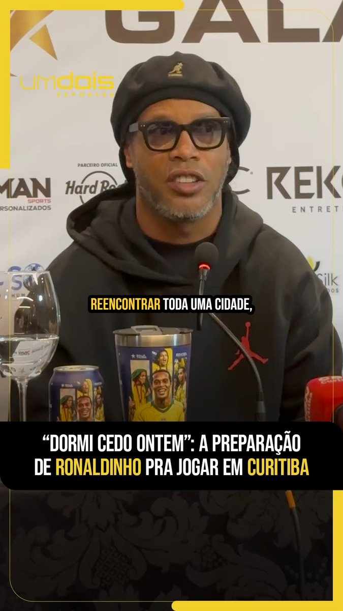 Ronaldinho, wearing a black Kangol cap, glasses, and a black Jordan hoodie, speaks at a press conference for the "Galácticos Game," with promotional cups of his image visible on the table. Subtitles read "REENCONTRAR TODA UMA CIDADE," and the caption below states "'DORMI CEDO ONTEM': A PREPARAÇÃO DE RONALDINHO PRA JOGAR EM CURITIBA."