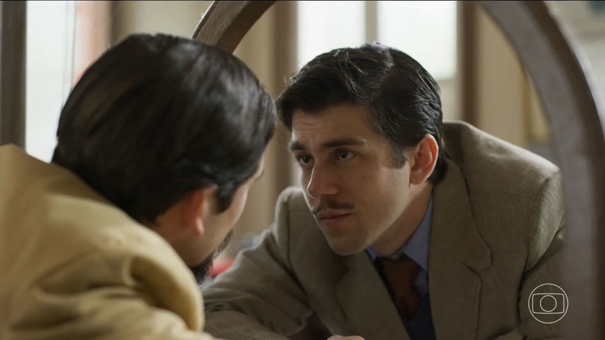 A scene from a Brazilian telenovela (Globo logo visible) showing a man with dark hair in a suit looking intently at another man reflected in or facing him through a circular frame.
