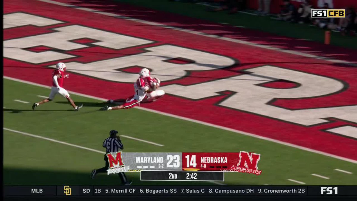 The image shows a Nebraska Cornhuskers football game on FS1, with the on-screen scoreboard displaying Maryland leading Nebraska 23-14 with 2:42 remaining in the 2nd quarter, as a player secures a catch in the red end zone painted with "HUSKERS" lettering. This relates to the social media post promoting a viewer pick for "Dwayne McDougle III" watching the @HuskerFootball game on FS1.