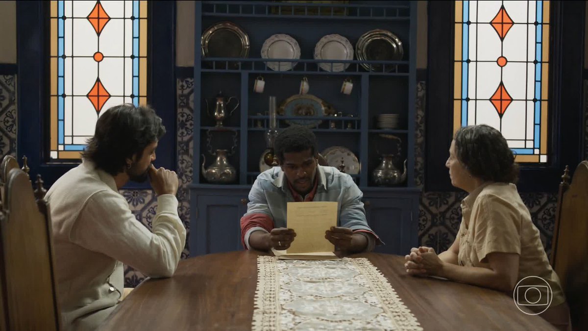 Three people sit at a dining table in a room featuring stained glass windows and a blue cabinet with decorative plates; the man in the center is reading a document.