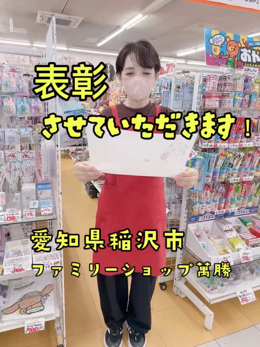 A store employee wearing a mask and a red apron holds up a drawing in a Japanese retail shop, with overlaid text reading "表彰させていただきます！", "愛知県稲沢市", and "ファミリーショップ萬脇".