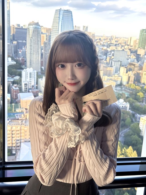 A young woman with long brown hair poses indoors holding a ticket in front of a large window overlooking a cityscape.