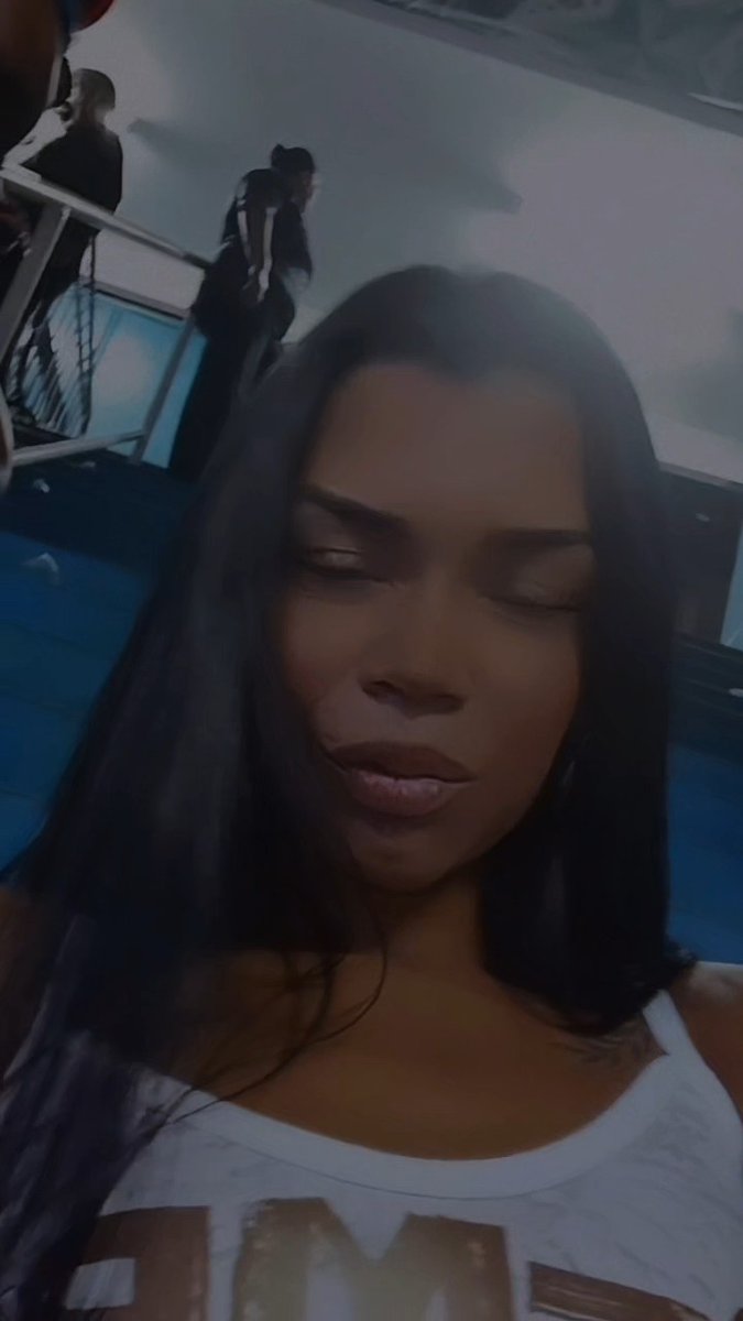 A woman with long dark hair and a white top looks downward with her eyes nearly closed, with blurred figures visible in the background.