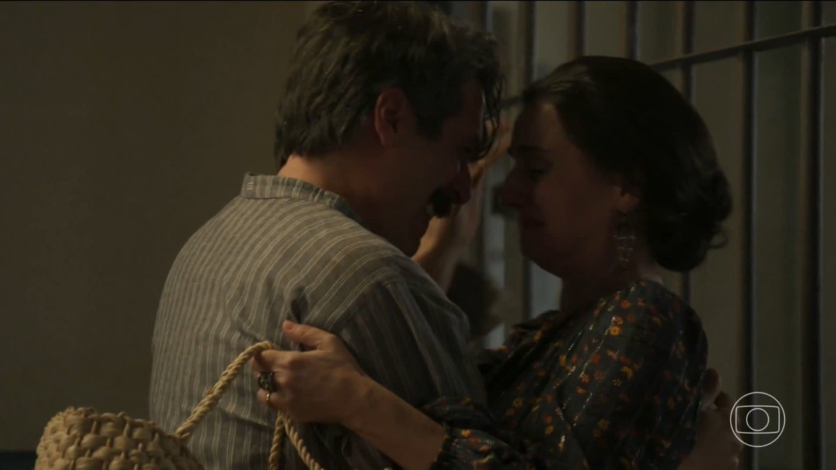 In a dimly lit scene that appears to be from a TV show, a man and a woman embrace closely, foreheads touching, with the woman holding a woven straw bag, against a backdrop of prison bars.