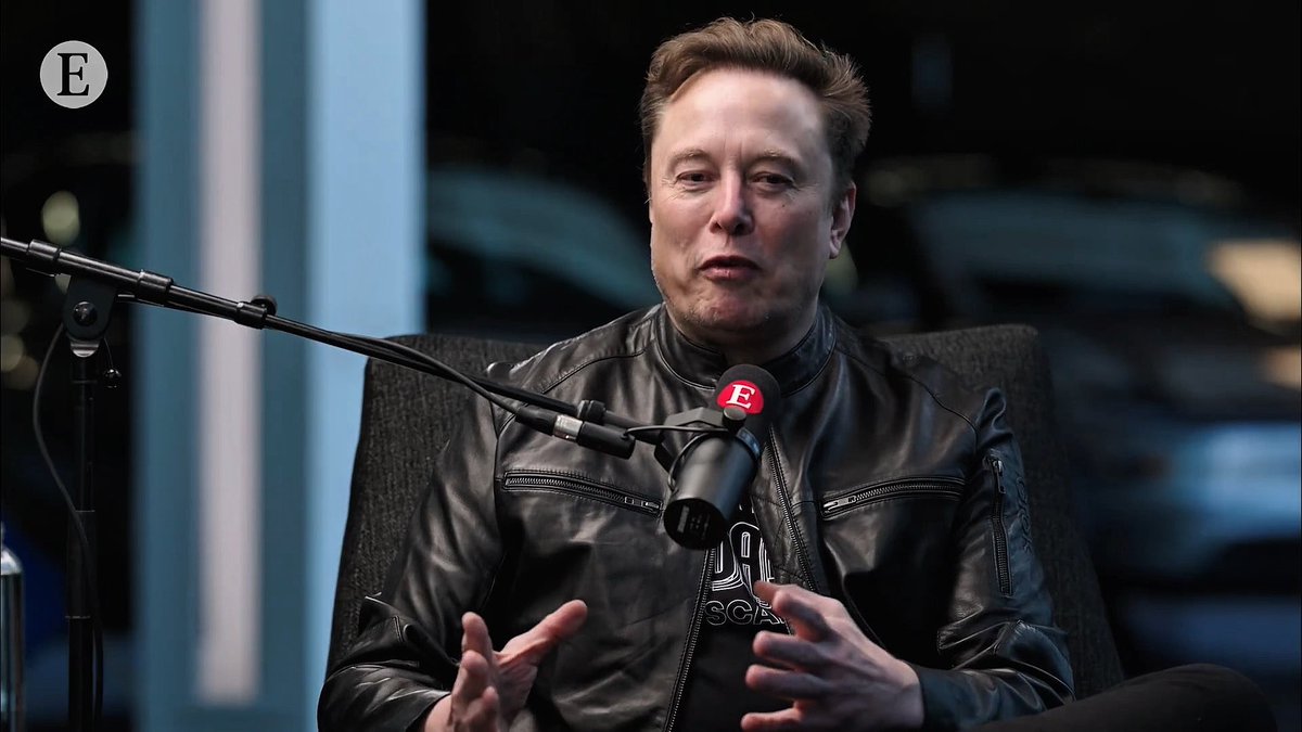 Elon Musk speaks into a microphone while wearing a black leather jacket, seated in what appears to be a studio setting with an 'E' logo visible.