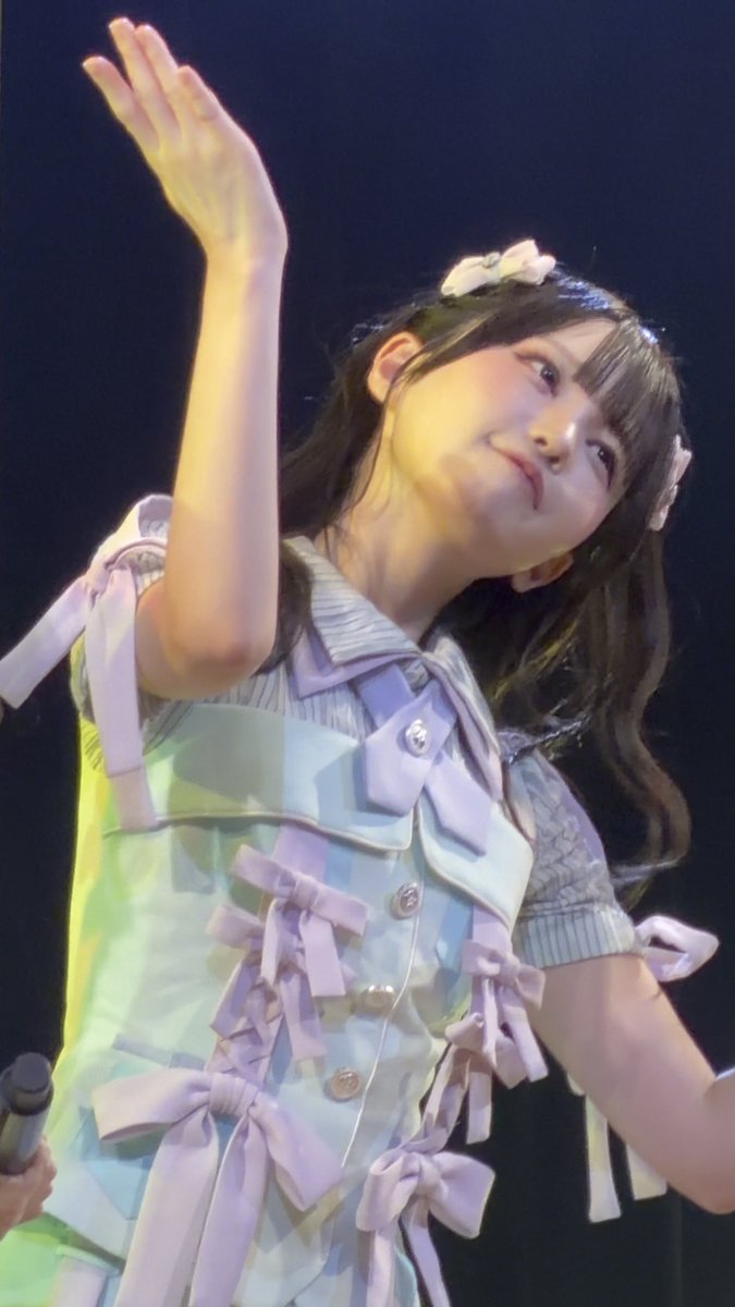 A young woman on stage with her arm raised, wearing a pastel checkered outfit featuring bows and hair clips.